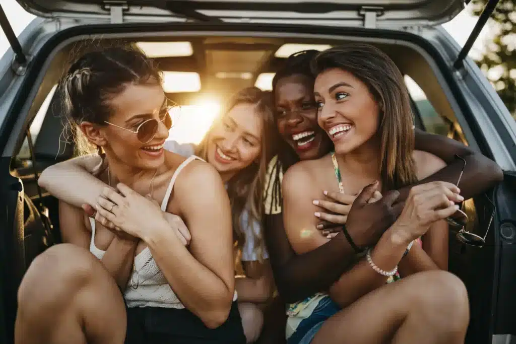 Groupe d'amies diverses riant joyeusement ensemble à l'arrière d'une voiture, au coucher du soleil, symbolisant la résonance émotionnelle.