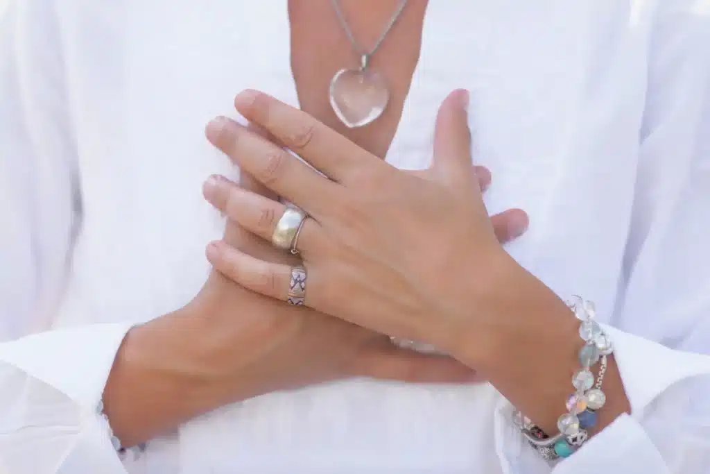 Gros plan sur les mains d'une femme posées sur son cœur, portant un collier en cristal, illustrant la connexion à son énergie vitale et son taux vibratoire.