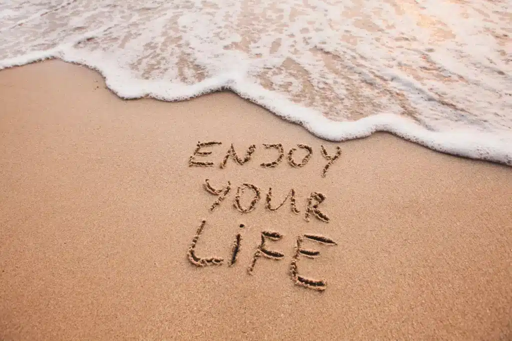 Les mots "Enjoy Your Life" écrits dans le sable sur une plage, avec une vague douce arrivant, symbolisant le pouvoir des pensées et émotions positives.