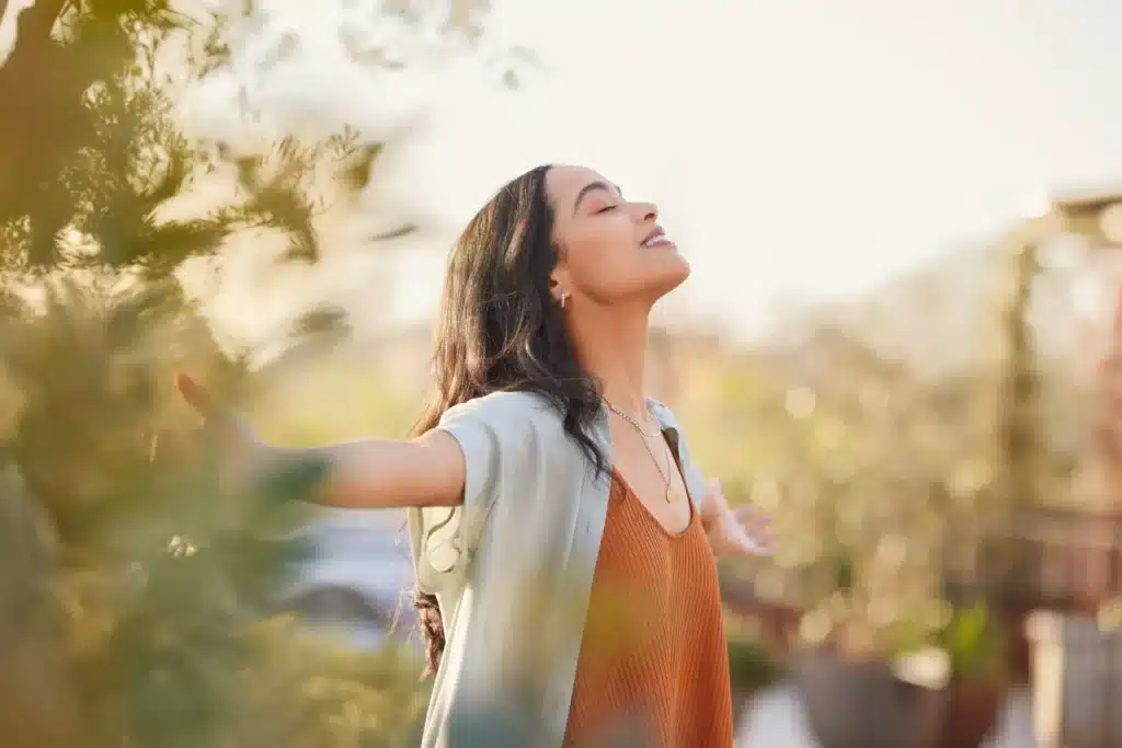Femme sereine, les yeux fermés et les bras ouverts, baignant dans la lumière du soleil, symbolisant l'harmonisation avec l'énergie de la nature pour élever son taux vibratoire.